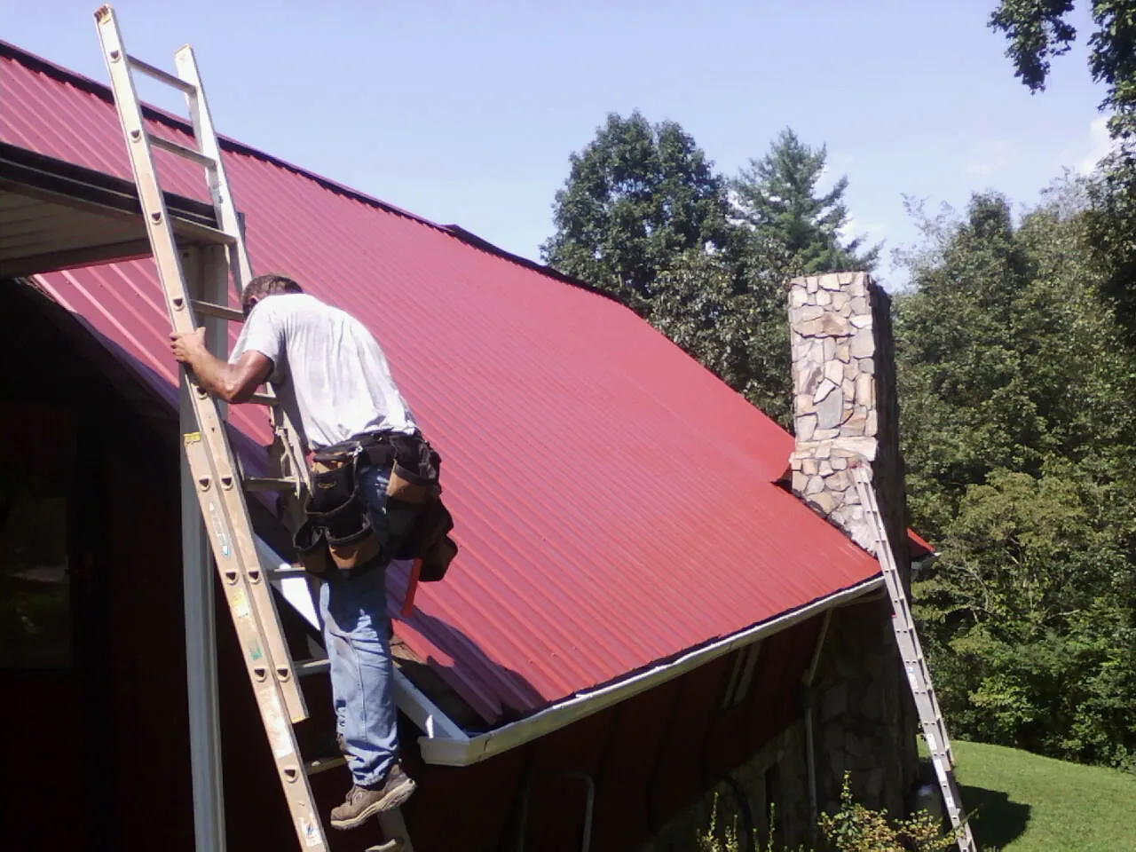 Roofer on a ladder beside a red metal roof and stone chimney — Soffit & Fascia Repair by Roofing Brothers in Dakota City